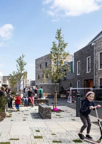 Children and adults gather and play along a paved walkway lined with young trees, with modern brick buildings on one side and a clear blue sky overhead.
