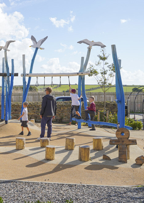 Children playing on a blue playground structure with a swing, climbing elements, and a seesaw, with a child in a white shirt and red hat swinging, others standing or walking nearby, on a sandy surface with wooden log obstacles in the foreground. A wooden fence, trees, and a partly cloudy sky are in the background.