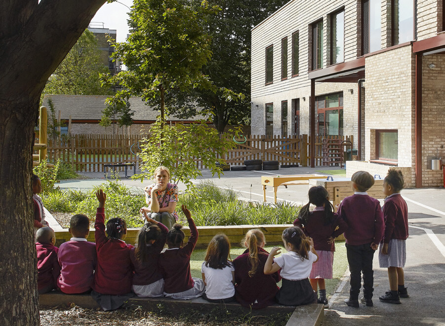 A group of young students, mostly wearing maroon uniforms, sit and stand outdoors in a schoolyard, paying attention to a woman with a bob haircut and floral shirt who is sitting on a low wall. Some students have their hands raised, and a few girls are standing on the right. The background includes trees, a wooden fence, and a modern building with large windows.