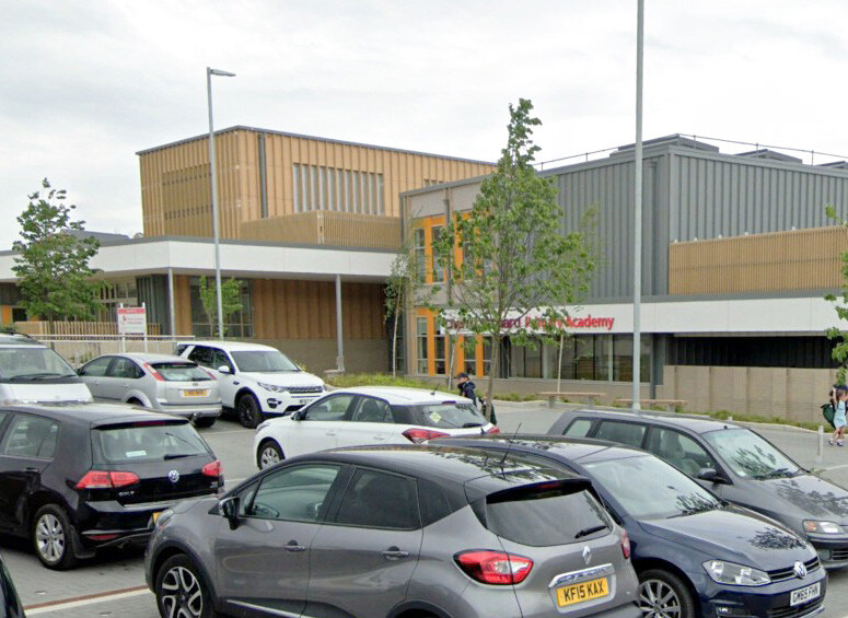 Multi-story building with wooden and metal exterior panels, large windows, and a sign reading "Academy" above the entrance. Parking lot in foreground with various cars and a few pedestrians walking past.