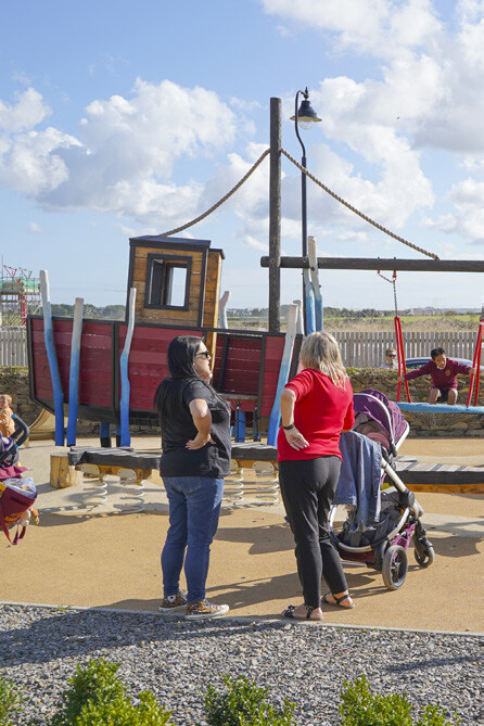 Two women standing and talking near a stroller, with children playing on a pirate ship-themed playground structure and a swing set in the background under a partly cloudy sky.