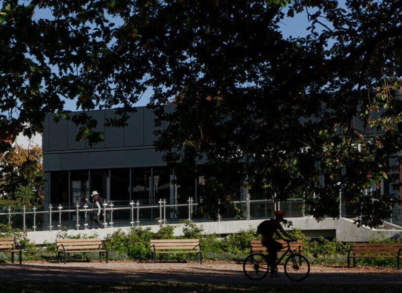 A person wearing a red helmet riding a bicycle along a path with wooden benches and bushes, partially shaded by large tree branches overhead, in front of a modern building with large glass windows.