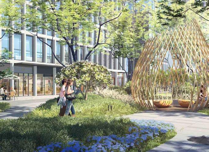 Two women walk and talk near a small tree in a landscaped urban park with green shrubs and blue flowers, alongside a modern glass building, with a large wooden oval-shaped sculpture with open slats and seating inside.