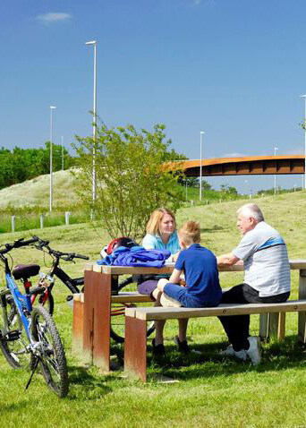 A man and two children sitting on a wooden bench outdoors, with bikes parked nearby. One child has red hair and the other blonde, and the man has gray hair. They are engaged in conversation on a grassy area with a tree, a bridge in the background, and a blue sky.