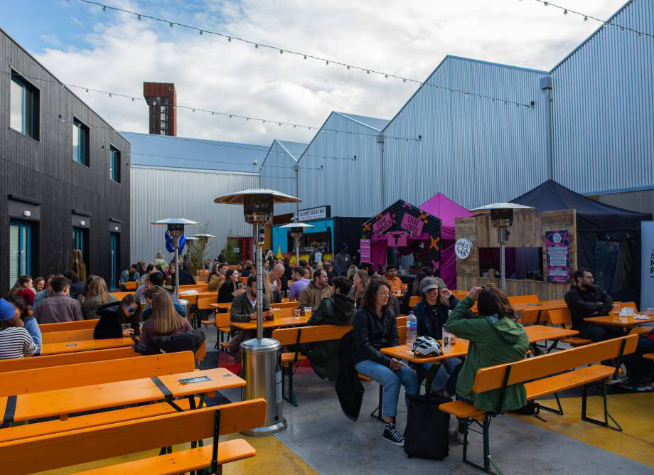 Crowd of people sitting at orange benches under string lights in an outdoor urban space with industrial buildings, some tents, and a purple and black stage backdrop.
