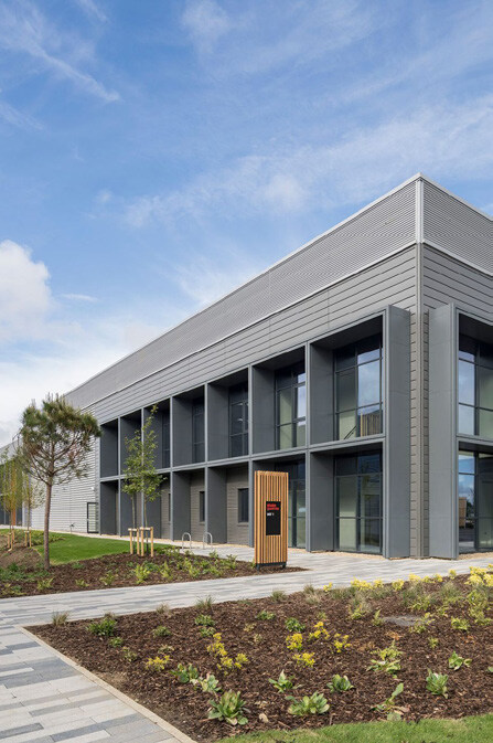 A modern two-story building with gray exterior panels and large glass windows, surrounded by a landscaped area with young trees, a paved walkway, and a wooden information kiosk, under a partly cloudy sky.