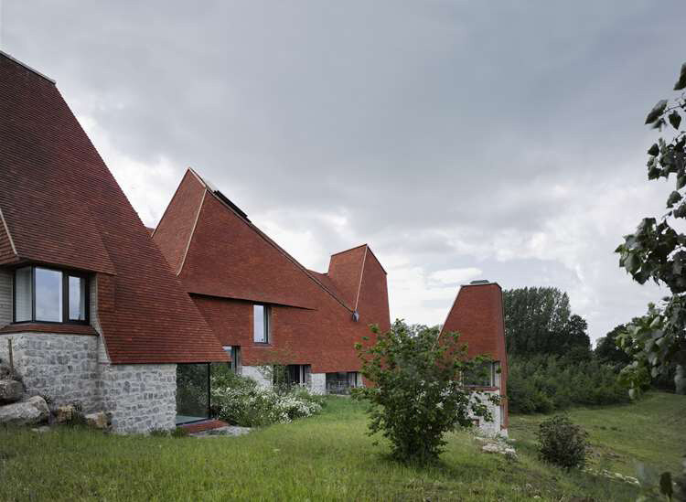 Multiple angular red brick roofs with steep slopes and sharp edges, some over stone walls, under a cloudy sky, with green grass and trees in the surroundings.