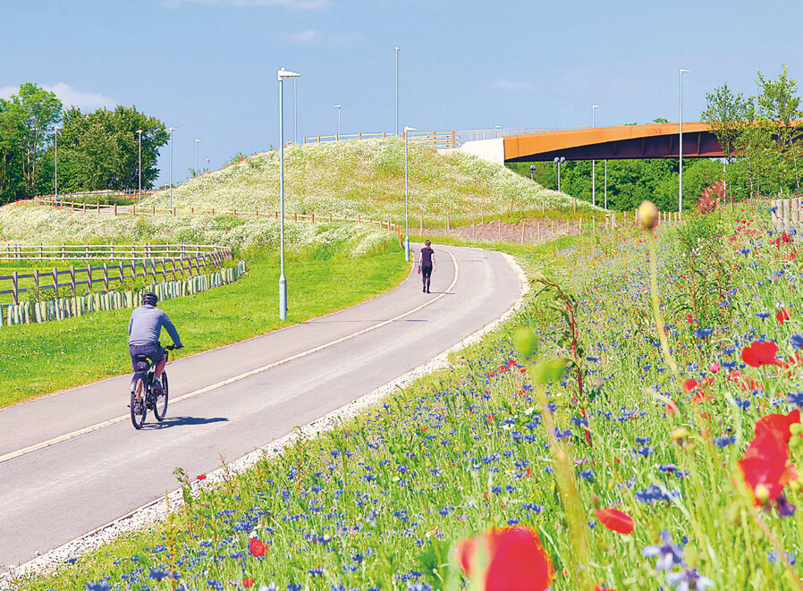 A winding paved path through a grassy hillside with wildflowers, a cyclist rides downhill, a person walks uphill, and a bridge spans above in the background, under a partly cloudy sky.