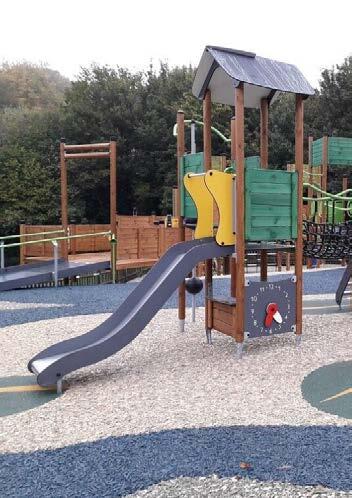A children's playground featuring a slide with a yellow dolphin-shaped handle, a small covered platform with a roof, surrounded by wooden fencing, trees in the background, and a gravel ground surface.