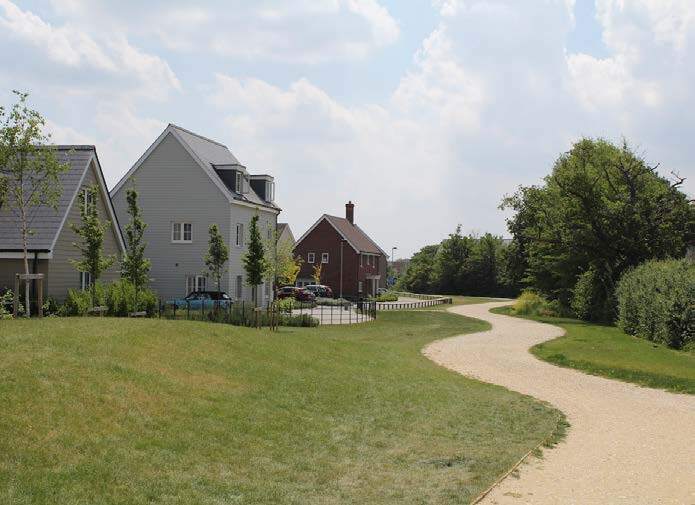 A paved pathway curves through a grassy area with trees and bushes, with modern multi-story houses in the background under a partly cloudy sky.