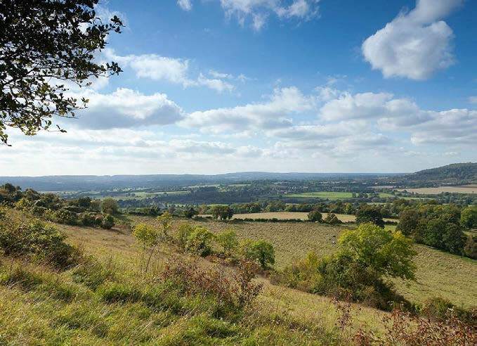 Gentle hillside with green grass and scattered trees, overlooking a patchwork of fields and distant rolling hills under a partly cloudy blue sky.