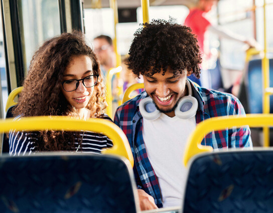 Two passengers seated next to each other on a bus; the person on the left has curly hair and wears a striped shirt, the person on the right has curly hair and wears a plaid shirt over a white t-shirt with headphones around their neck.