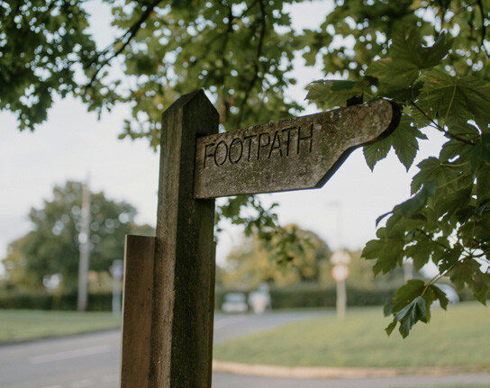 Wooden footpath sign with faded, weathered lettering, mounted on a post among leafy branches; sidewalk and street visible in the background.