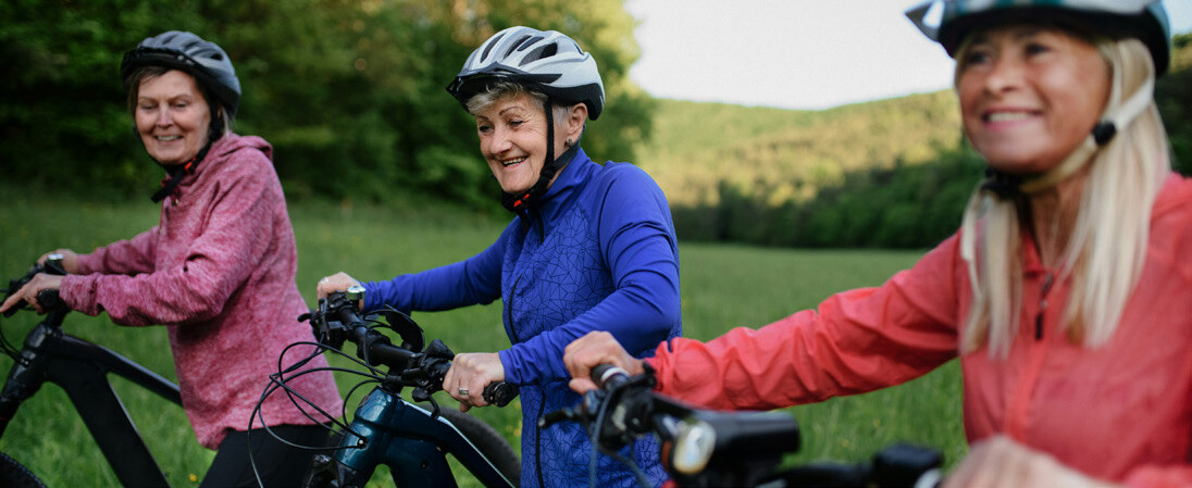 Three women wearing helmets and outdoor jackets ride bicycles through a grassy field with trees and hills in the background.