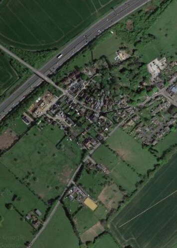 A rural village with densely packed houses connected by narrow winding roads, surrounded by green fields and pastures, with a main highway running along the top edge and a canal crossing the lower part of the image.