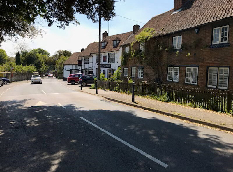 A quiet street with parked cars along the curb, a white car driving away, brick and white houses with multiple windows and pitched roofs, some greenery and vines on the buildings, and shadows of trees cast on the road and sidewalk.