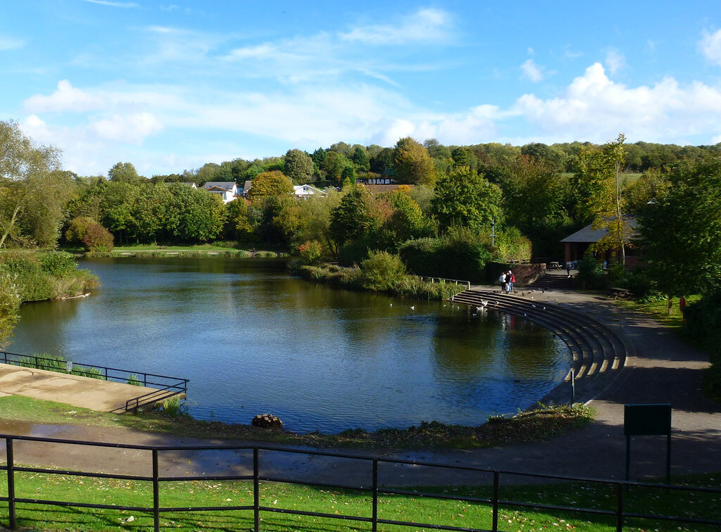 A small lake surrounded by trees and greenery with a curved, tiered concrete seating area on the right and a paved pathway. Two people walk along the path, and some birds are on the water. Houses and a hill are visible in the background under a blue sky with scattered clouds.