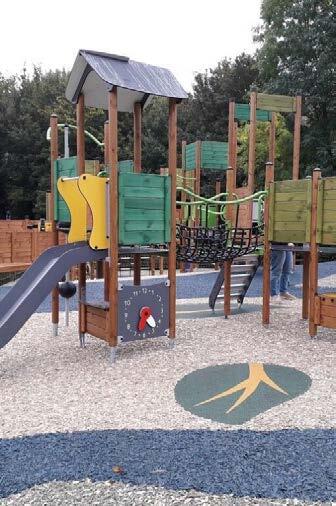 A colorful wooden playground structure with a slide, bridges, and climbing elements, set on gravel with a painted tree trunk design on the ground, surrounded by trees and a fence.