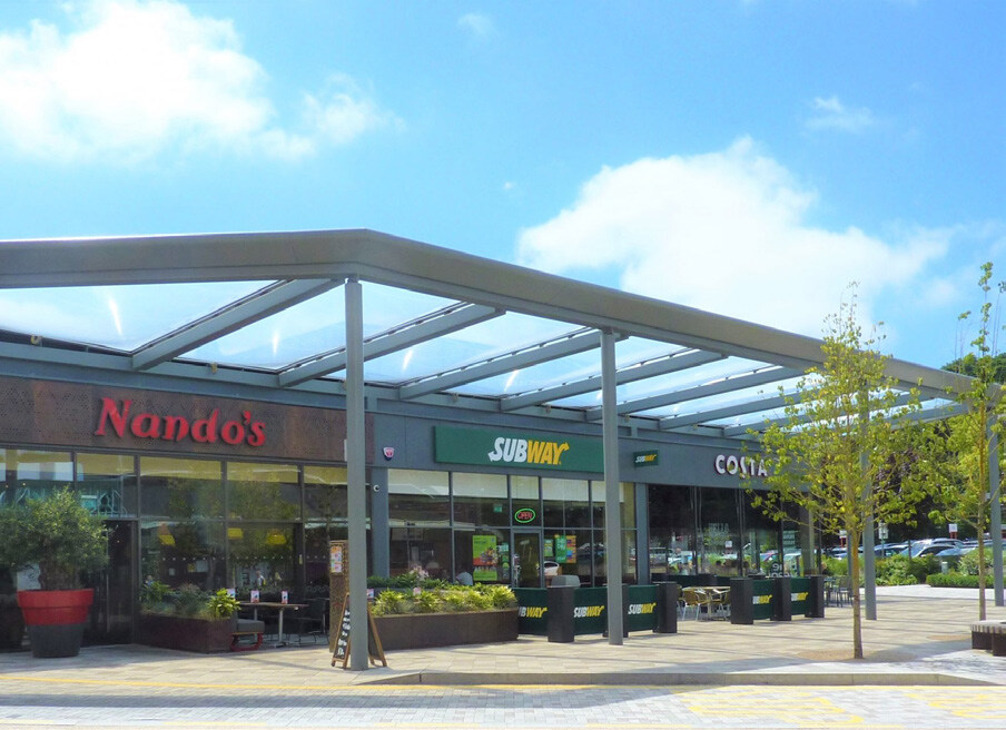 Nando's, Subway, and Costa Coffee storefronts under a glass canopy, outdoor seating area with tables and chairs, young tree with green leaves, parking lot with cars in the background, blue sky with white clouds.