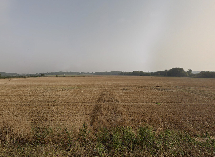 Vast, flat farmland with dry, harvested crops, bordered by green bushes in the foreground and a line of trees on the horizon under a cloudy sky.