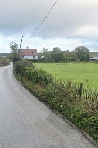 Gravel country road curves left alongside a green grassy field with bushes, leading toward a white house with a red roof and multiple chimneys. Power lines run parallel to the road. Overcast sky.
