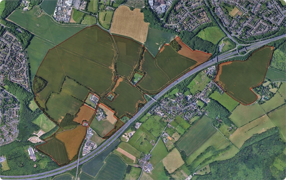 Aerial view of a landscape with a highway crossing diagonally, surrounded by green fields, patches of woodland, and residential neighborhoods. Fields are outlined with red borders.
