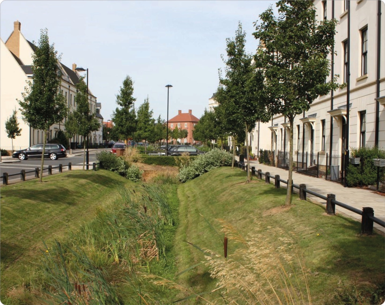 A landscaped urban area with a narrow, grassy drainage channel in the center, bordered by mowed grassy ridges. On the right, there is a sidewalk lined with young trees and black railings in front of multi-story residential buildings with beige facades and numerous windows. On the left, a street with parked cars and additional trees extends into the distance under a partly cloudy sky.