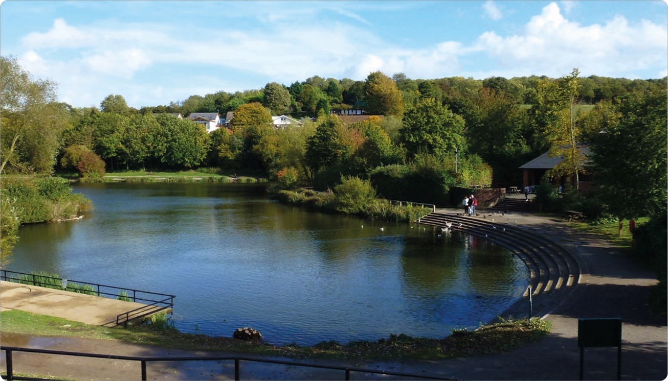 A river with gentle curves, lined with trees in various shades of green and orange, reflecting the sky. On the right side, tiered concrete steps lead to the water, with a small group of people walking nearby. In the background, houses are nestled in a hillside covered with trees under a partly cloudy blue sky.