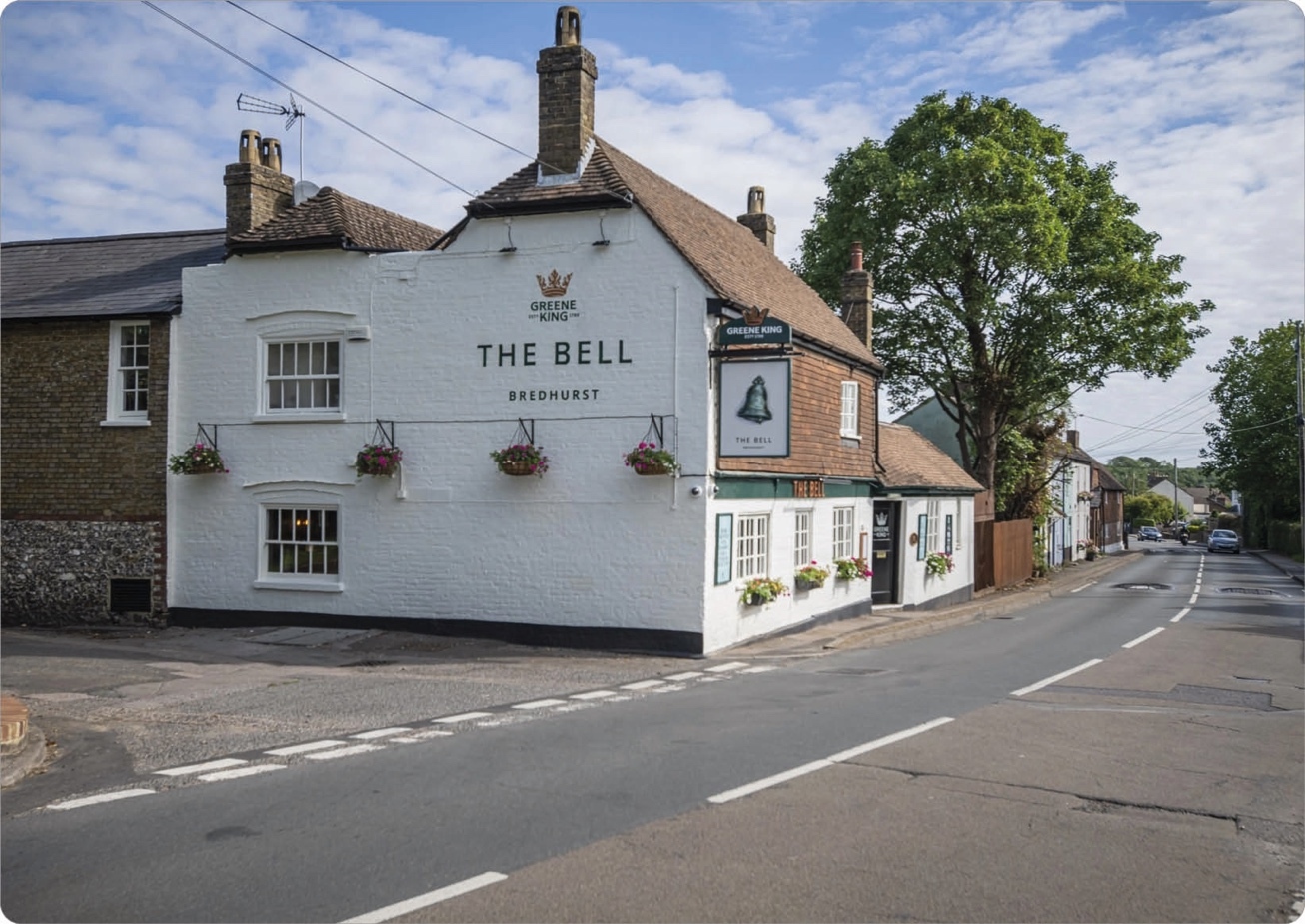 A white-painted, two-story pub named "The Bell" in Bredhurst, with flower baskets hanging from the front and a sign featuring a bell. The building has a sloped, tiled roof with brick chimneys and is situated on a corner street with trees and neighboring houses visible.
