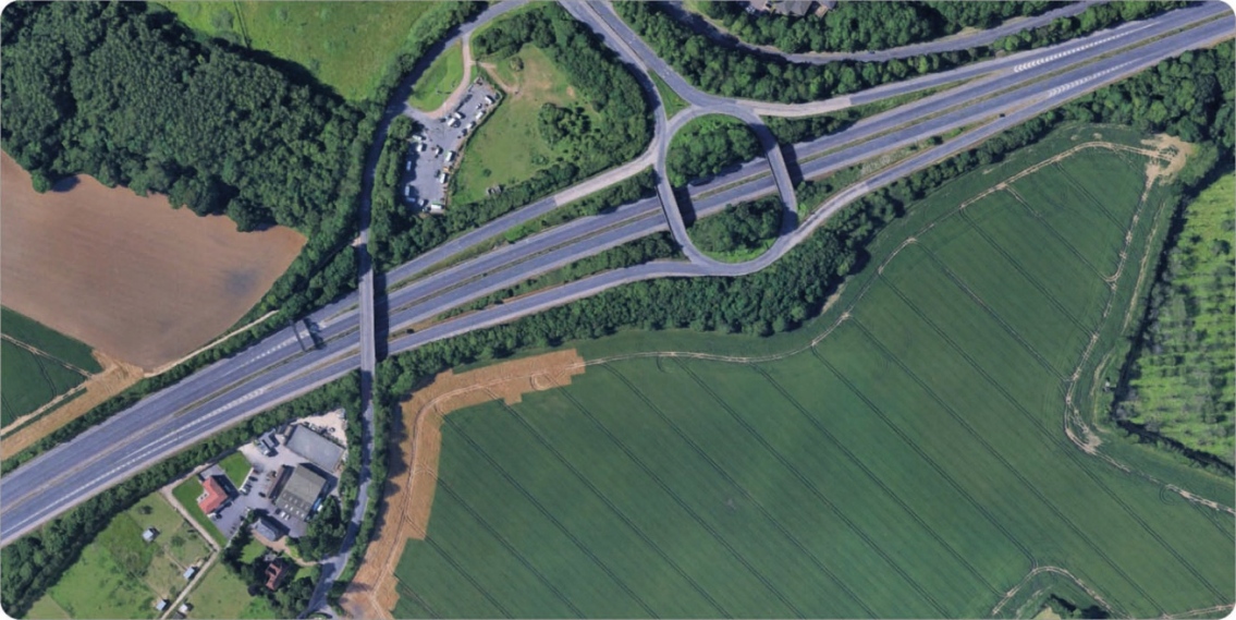 Aerial view of a highway interchange with multiple overpasses, surrounded by green fields and patches of woodland.