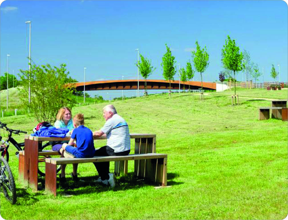 A family of three with a bicycle sitting on a wooden bench in a grassy park, with a bridge and trees in the background on a sunny day.