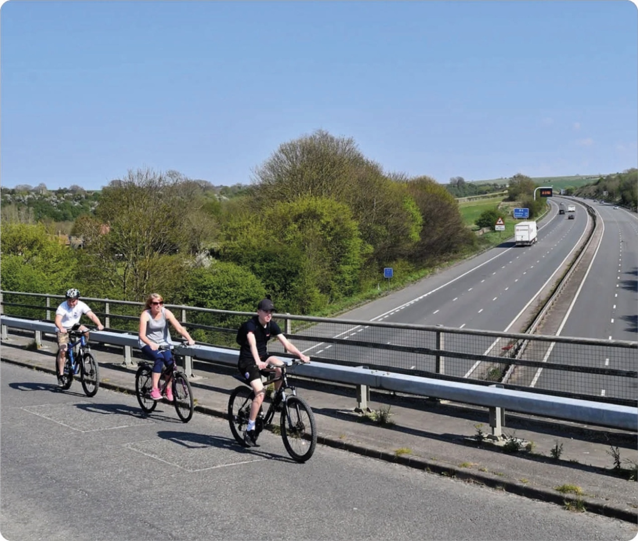 Three children riding bikes on a roadside path with a metal guardrail, overlooking a highway with moving vehicles, green trees, and blue sky in the background.