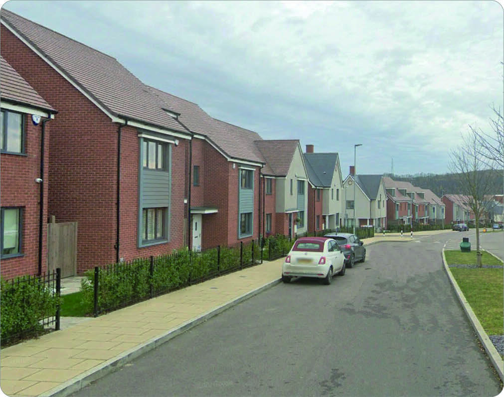 Row of modern, two-story houses with brick and light-colored siding, sloped roofs, and small front yards. Cars parked along the street, which curves gently. Overcast sky with light cloud cover.
