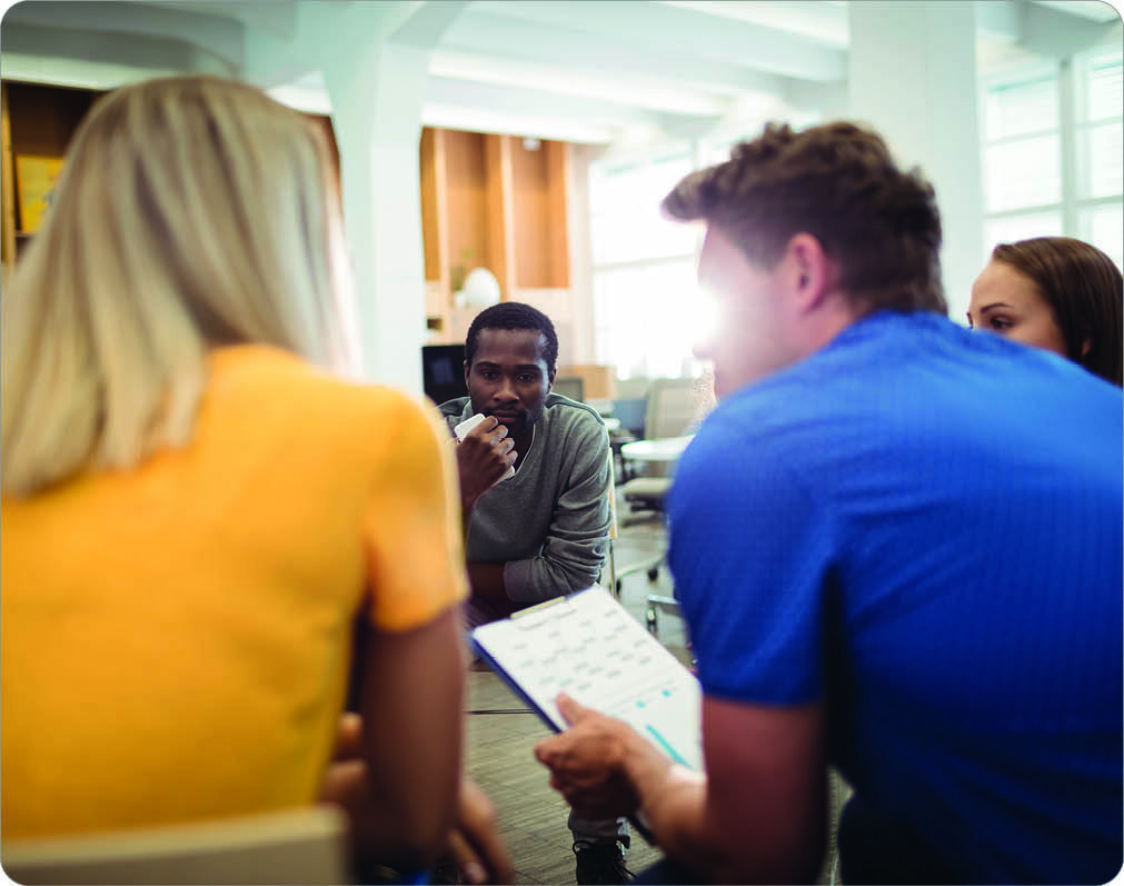 A group of four diverse people sitting in a circle in a bright, modern room, engaged in a discussion. The man in a blue shirt is holding a clipboard with notes, while the others listen attentively.