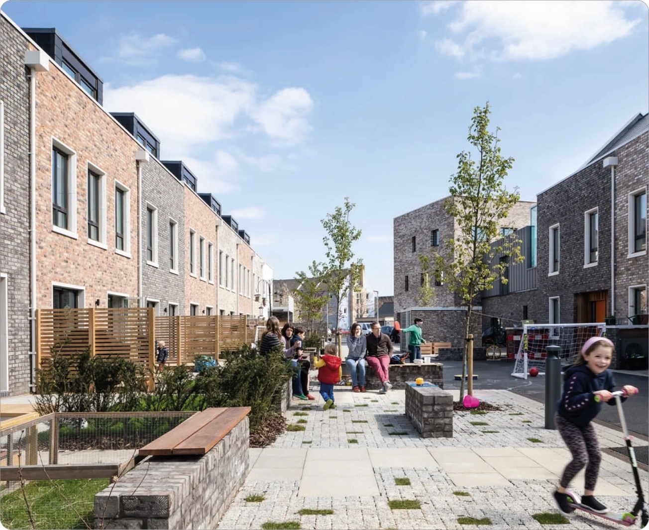 Children and adults sitting on brick benches and standing around, some playing with balls, in a courtyard with young trees and paved walkways, surrounded by modern townhouses with brick facades under a blue sky with scattered clouds.