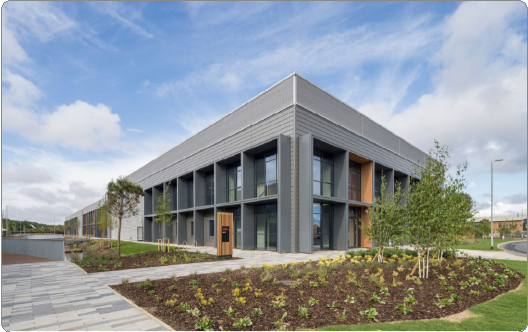 Modern two-story building with large glass windows, gray metal exterior, surrounded by landscaped garden, small trees, and a paved walkway under a partly cloudy sky.