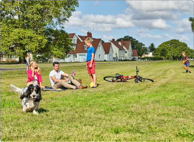 Girl with long blonde hair in a pink jacket sits on the grass, holding a phone to her ear. A man with a face visible, wearing a white t-shirt and beige pants, is sitting on the grass next to a girl with long blonde hair in a pink jacket, drinking from a bottle. A boy in a blue shirt and red shorts stands nearby, looking down. A red bicycle lies on its side on the grass. A small black and white dog runs toward the camera. In the background, a girl in a colorful dress runs across the field. There are houses with pitched roofs and trees under a partly cloudy sky.