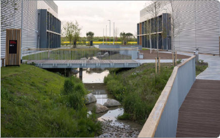 Two modern buildings with metallic exteriors flank a small bridge over a narrow creek, with a wooden walkway and green grass on both sides.