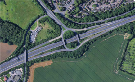 A highway interchange with multiple lanes, overpasses, and surrounding green fields and trees.