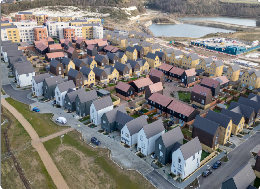 Aerial view of a residential neighborhood with closely spaced modern houses, some with white or dark exteriors, pitched roofs, and small gardens. In the background, larger apartment buildings, a body of water, and construction or storage area are visible.