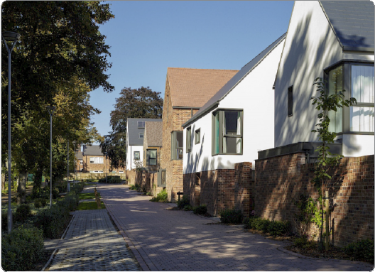 White and brick houses with angled roofs line a paved sidewalk, trees cast shadows, and a clear blue sky is above.