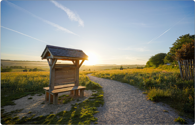 A wooden bench with a small roof on a dirt path in a grassy field at sunset, with trees and a wooden fence in the background.